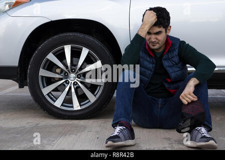depressed man sitting on a ground leaning on his broken car. Sad, angry and frustrated person. Emergency. Stock Photo