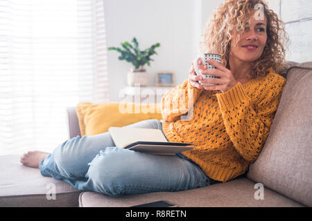 Beautiful 40 years old caucasian lady sit down on the sofa drinking tea and reading a book for afternoon indoor leisure activity at home - after work  Stock Photo