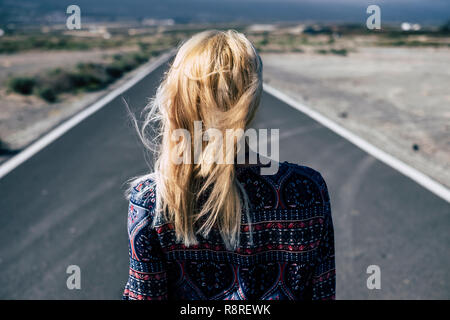 Blonde young girl viewed from rear walk on a long straight road in the country side. Decisions and future concept for millennial people looking for a  Stock Photo
