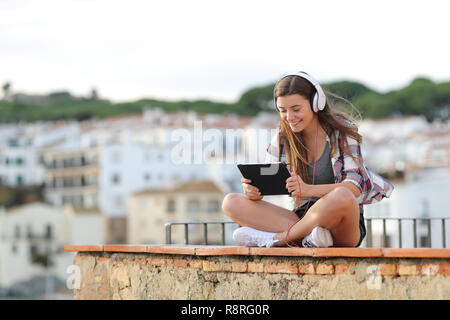 Happy teen wearing headphones watching and listening online video on a tablet sitting on a ledge on vacation Stock Photo