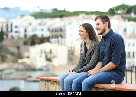 Happy couple or friends looking away on vacation sitting on a ledge in a coast town Stock Photo