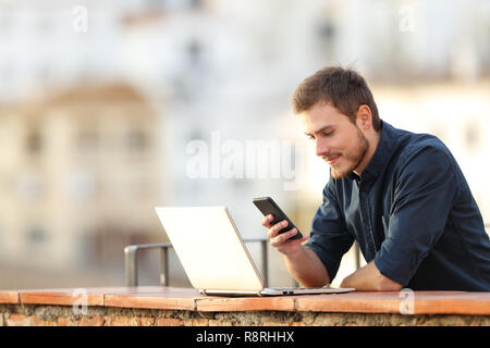 Serious man with a laptop checking smart phone content in a balcony in a town Stock Photo