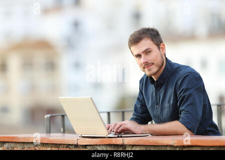 Confident man using a laptop and looking at camera in a balcony in a town Stock Photo