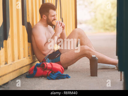 Man eats a hamburger after a workout. Very hungry, fatty and unhealthy food. Stock Photo