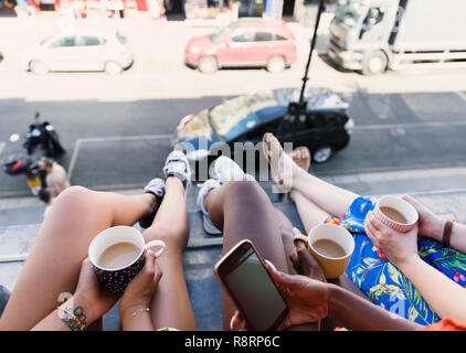 Women friends drinking coffee, dangling legs out urban apartment window Stock Photo