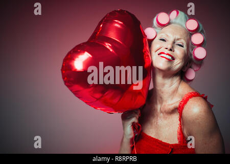Portrait playful senior woman with hair in curlers holding heart-shape balloon Stock Photo