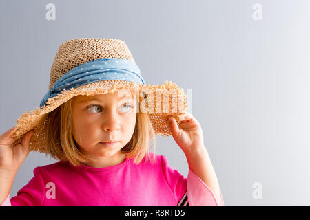 Young pretty girl in pink dress and rural straw hat looking away to the side of copy space on plain grey wall background, while holding her hat with b Stock Photo