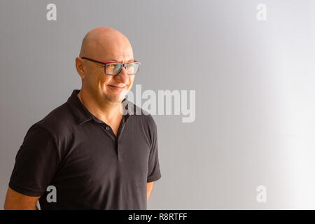 Adult bold man in glasses and black polo half turn half-length portrait, looking to the side of grey copy space and smiling Stock Photo