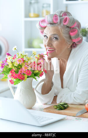 Portrait of senior woman in bathrobe with curlers sitting at table with laptop Stock Photo