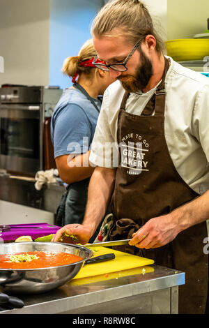 Male Chef Slicing Avocado for  Wedding Meal - Kitchen Set, Text on Apron "Made to go presents, Great Bakery, Enjoy" Stock Photo