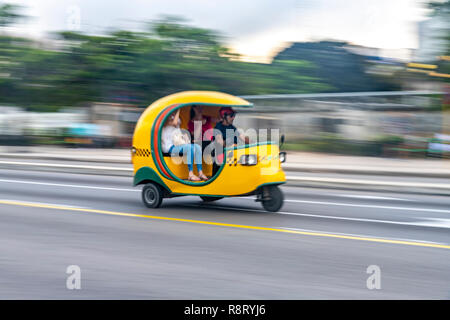 Cuban man driving a speeding bright yellow Coco Taxi with two passengers in Havana Cuba Stock Photo