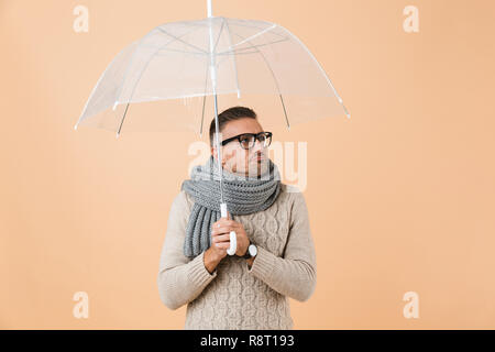 Portrait of a cold man dressed in sweater and scarf standing under umbrella isolated over beige background