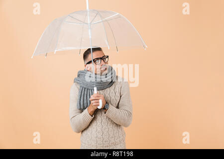 Portrait of a sad man dressed in sweater and scarf standing under umbrella isolated over beige background