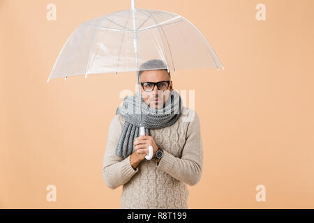 Portrait of a confused man dressed in sweater and scarf standing under umbrella isolated over beige background