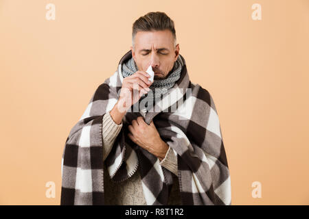Portrait of a sick man wrapped in a blanket standing isolated over beige background, having runny nose, using napkin Stock Photo