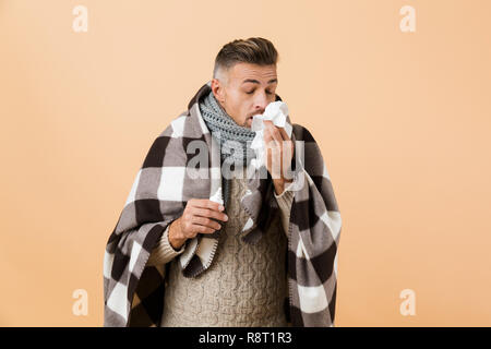 Portrait of a sick man wrapped in a blanket standing isolated over beige background, having runny nose, using napkin Stock Photo