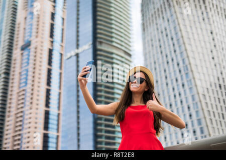 Portrait of young woman in red dress, sunglasses and summer hat make video call with thumbs up on the phone on downtown skycrapers background Stock Photo
