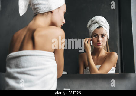 Pretty young woman with towel on head removing makeup with a cotton swab in front of mirror in bathroom Stock Photo