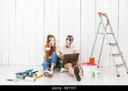 Photo of excited couple man and woman sitting on floor over white wall near ladder and using laptop while making renovation indoor Stock Photo