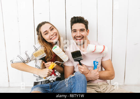 Image of excited couple man and woman holding paint rollers and brushes while standing over white wall during renovation Stock Photo