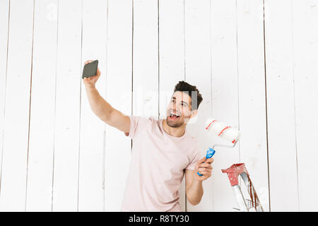 Portrait of young man 20s taking selfie on mobile phone while painting white wall with paint roller and making renovation Stock Photo