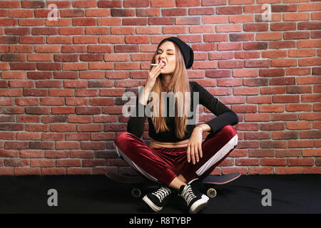 Photo of subcultural sporty girl 20s sitting on skateboard with cigarette against brick wall Stock Photo