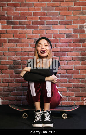 Image of teen sporty girl 20s laughing while sitting on skateboard against brick wall Stock Photo