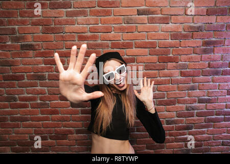 Image of stylish sporty girl in hat and sunglasses laughing while standing against brick wall Stock Photo