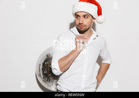Handsome young man wearing santa claus hat carrying silver disco ball isolated over white background Stock Photo