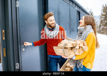 Young lovely couple dressed in colorful sweaters entering their modern home with firewoods in the mountains during the winter
