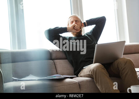Handsome young man working at home, sitting on a couch using laptop computer, yawning Stock Photo