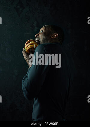 The young african american man eating hamburger and looking away on black studio background Stock Photo