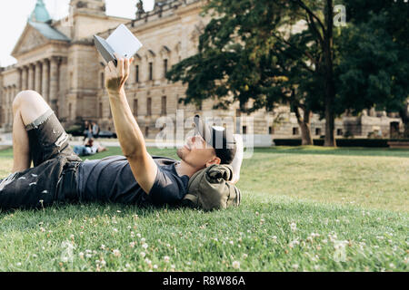 A young guy with a backpack or a student is lying on the grass during a break or is resting and using a tablet. He is watching a video or reading an e-book or something else. Stock Photo