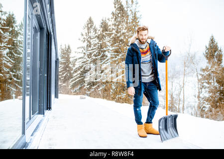 Portrait of a man in winter clothes standing with snow shovel on the terrace of the building in the mountains Stock Photo