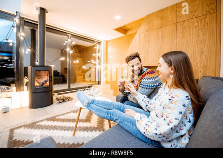 Young couple celebrating winter holidays sitting with wine on the couch in the beautiful decorated house with fireplace Stock Photo