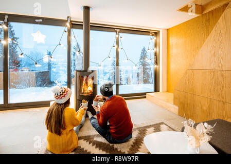 Young couple dressed in bright sweaters sitting near the fireplace in the modern house in the mountains durnig winter time Stock Photo