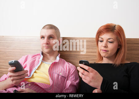 Young couple sitting on hotel bed using mobile phones and watching TV Stock Photo
