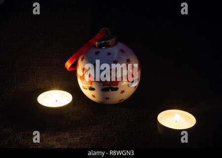 A decorative Christmas bauble depicting three Santa Clauses with two lit candles next to it against black background- Christmas concept Stock Photo