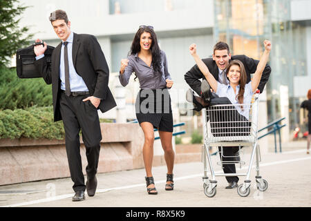 Group of young business people on the street near a supermarket in a hurry to sell, pushing their colleague in a shopping trolley. Stock Photo