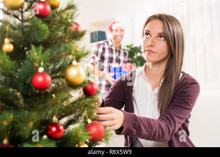 Young beautiful girl decorates a Christmas tree while her boyfriend holds a Christmas gift and he wants to surprise her. Stock Photo