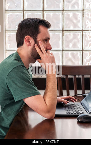 Young bearded man at home working remotely on laptop computer in front of glass block wall Stock Photo