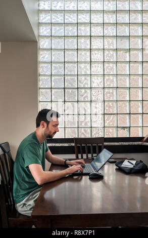 Young bearded man at home working remotely on laptop computer in front of glass block wall Stock Photo