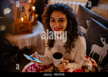 Beautiful smiling woman with curly hair is holding a smartphone and a cup of tea or coffee in her hands Stock Photo
