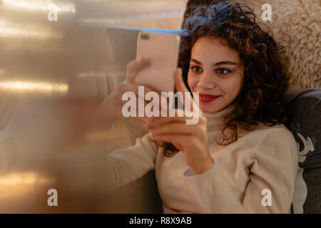 Beautiful young woman with curly hair is reading messages on her smartphone Stock Photo