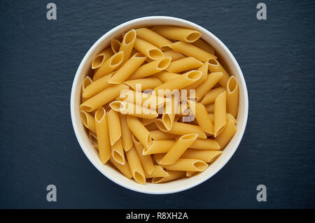 Pasta penne in white bowl on dark stone table. Top view. Raw penne pasta in a bowl. Stock Photo