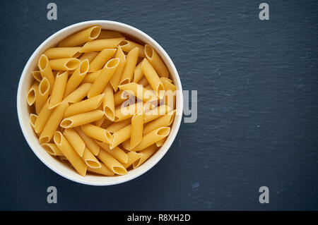 Pasta penne in white bowl on dark stone table. Top view. Raw penne pasta in a bowl. Stock Photo