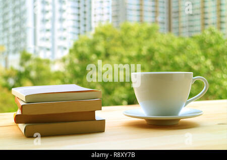 Stack of books and a cup of hot drinks on a table by the window with blurred green foliage and high buildings in background Stock Photo