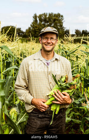 Smiling farmer standing in a corn field, holding bunch of maize cobs. Stock Photo