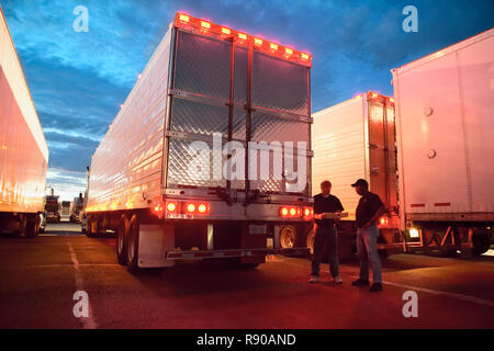 Two truck drives checking dispatch papers while standing next to truck trailers in a large parking lot at night. Stock Photo