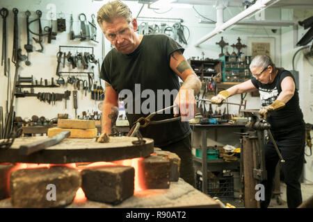 Caucasian male blacksmith working at a forge in the foreground and femal blacksmith at work in background. Stock Photo